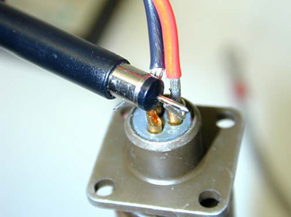Oscilloscope probe with its ground lead removed, using only the probe tip and barrel to bridge an output pin and its return pin directly on a circular interface connector